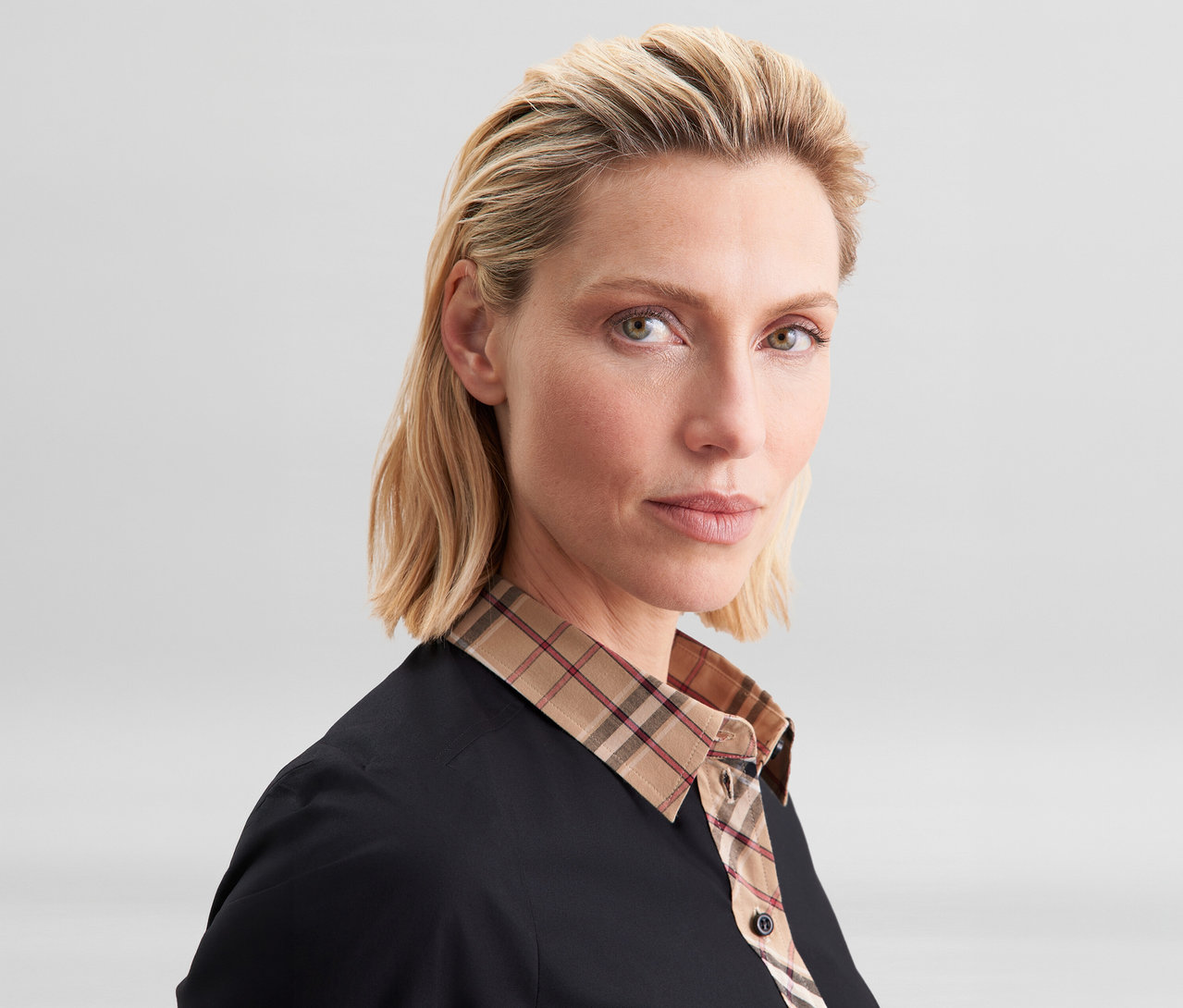 Blonde Frau trägt eine schwarze Seidensticker Popeline-Bluse mit Karodetail, die einen beigen, rot-schwarz karierten Kragen und eine Knopfleiste hat.