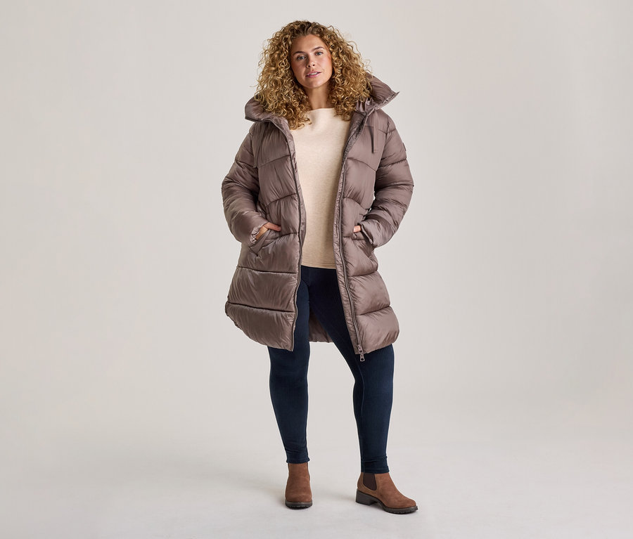 Frau mit lockigem Haar trägt einen langen braunen Steppmantel, ein helles Oberteil, dunkle Hose und braune Stiefeletten.