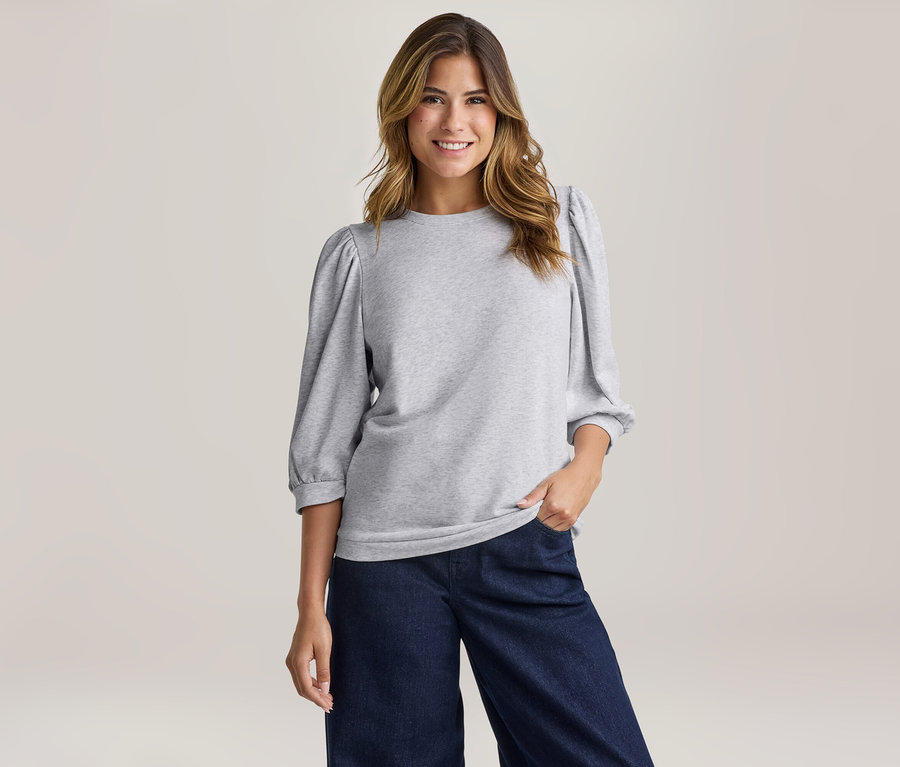 Eine lächelnde Frau mit langen, gewellten Haaren trägt ein hellgraues Sweatshirt mit Puffärmeln und eine dunkelblaue weite Hose.
