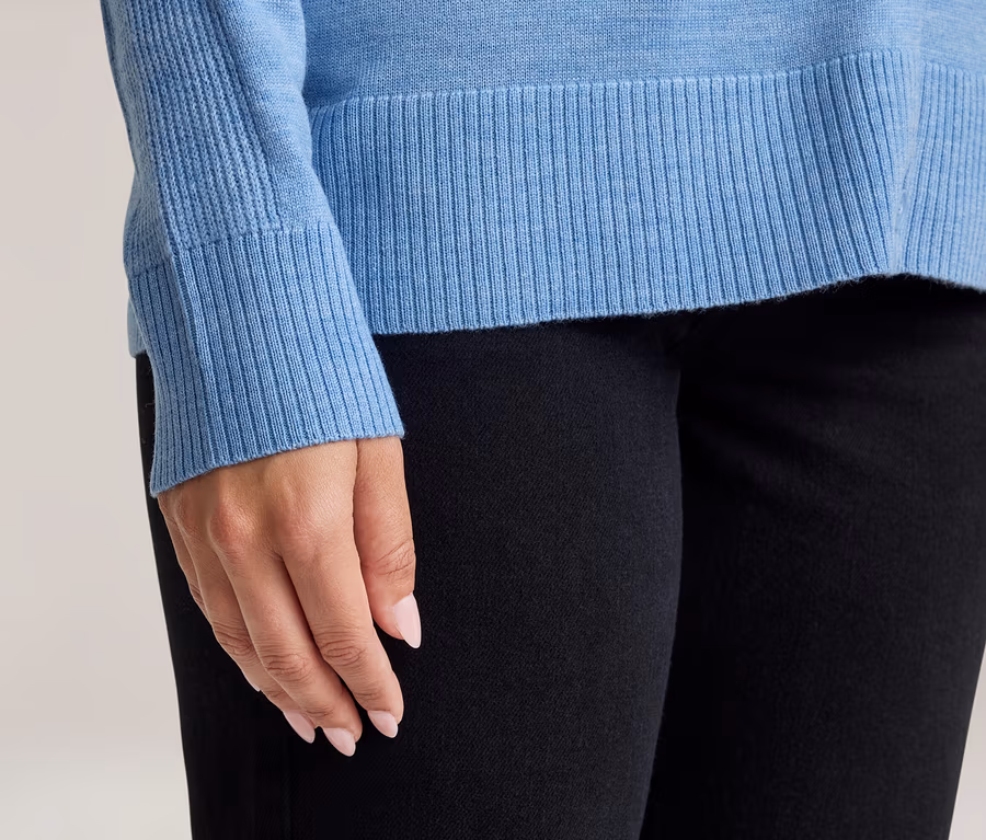 Nahaufnahme einer Person in einem hellblauen Pullover mit gerippten Bündchen und Saum sowie einer schwarzen Hose. Die Hände der Person mit hellrosa Nagellack sind sichtbar.