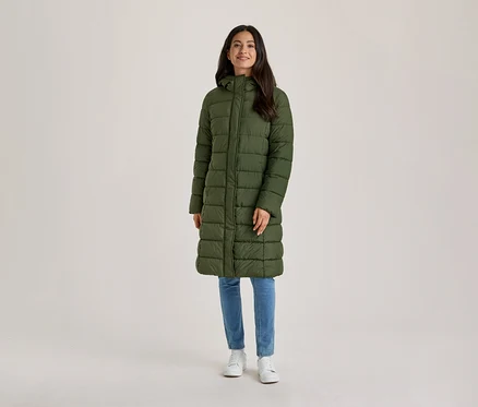 Eine lächelnde Frau mit dunklen Haaren steht frontal da und trägt einen langen, olivgrünen Steppmantel mit Kapuze, blaue Jeans und weiße Turnschuhe.