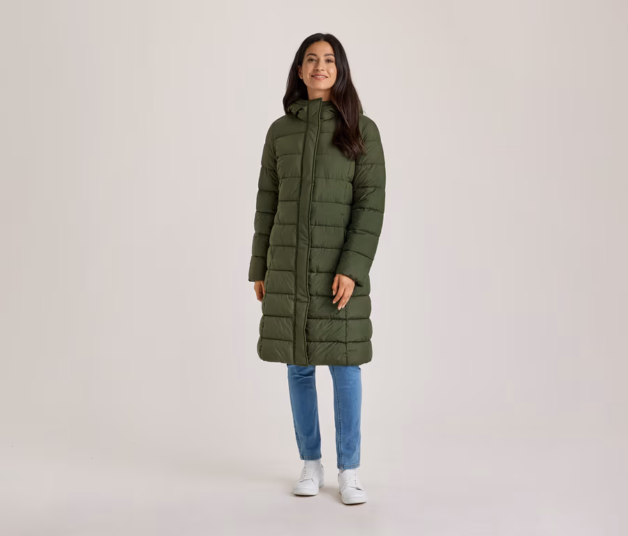 Eine lächelnde Frau mit dunklen Haaren steht frontal da und trägt einen langen, olivgrünen Steppmantel mit Kapuze, blaue Jeans und weiße Turnschuhe.
