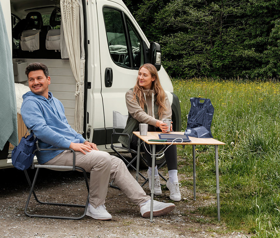 Ein Mann in einem hellblauen Sweat-Hoodie und eine Frau in einer beigen Sweatjacke und einer khakifarbenen Hybrid-Weste sitzen vor einem Campervan. Auf einem Tisch liegt eine dunkelblaue Pack-Weste.
