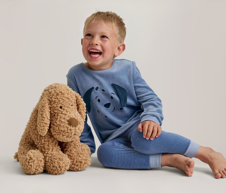 Ein lachender kleiner Junge in blauem Pyjama mit Hunde-Gesichtsmotiv sitzt neben einem flauschigen braunen Plüschhund.