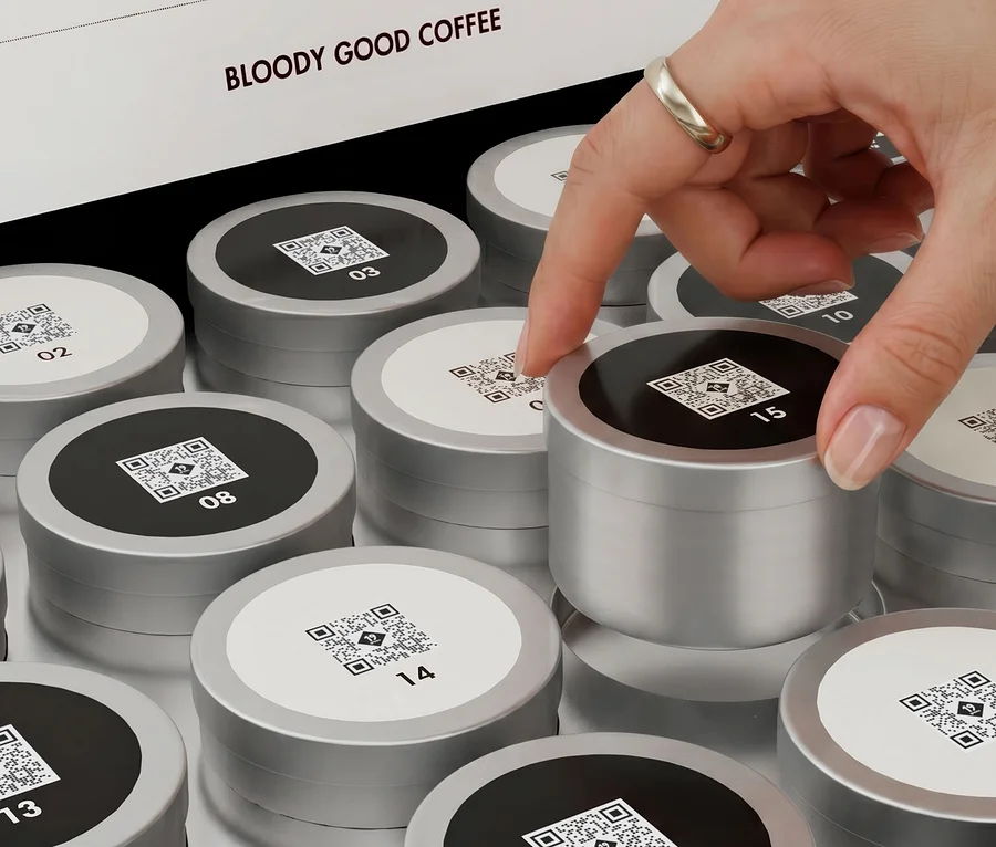 Eine Hand mit Ring nimmt eine silberne Kaffeekapsel mit schwarzer Oberseite und QR-Code aus einer Schachtel voller nummerierter Kapseln. Auf dem Deckel steht 'BLOODY GOOD COFFEE'.