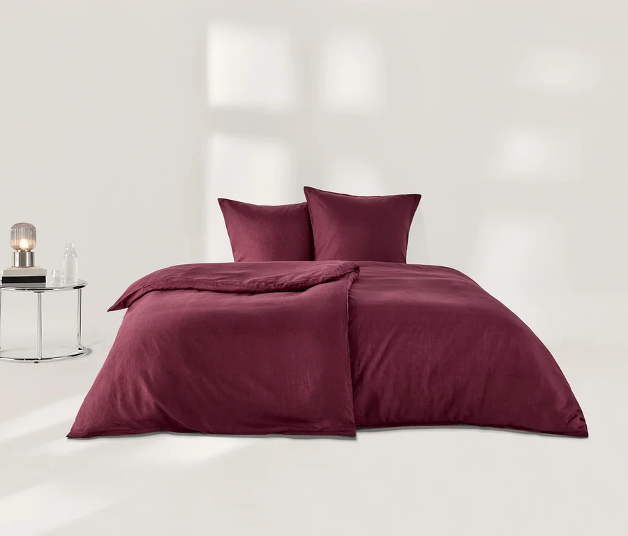 Beerenfarbene Leinen-Bettwäsche mit Bettbezug und zwei Kissen auf einem Bett, daneben ein Beistelltisch mit Lampe und Flasche.