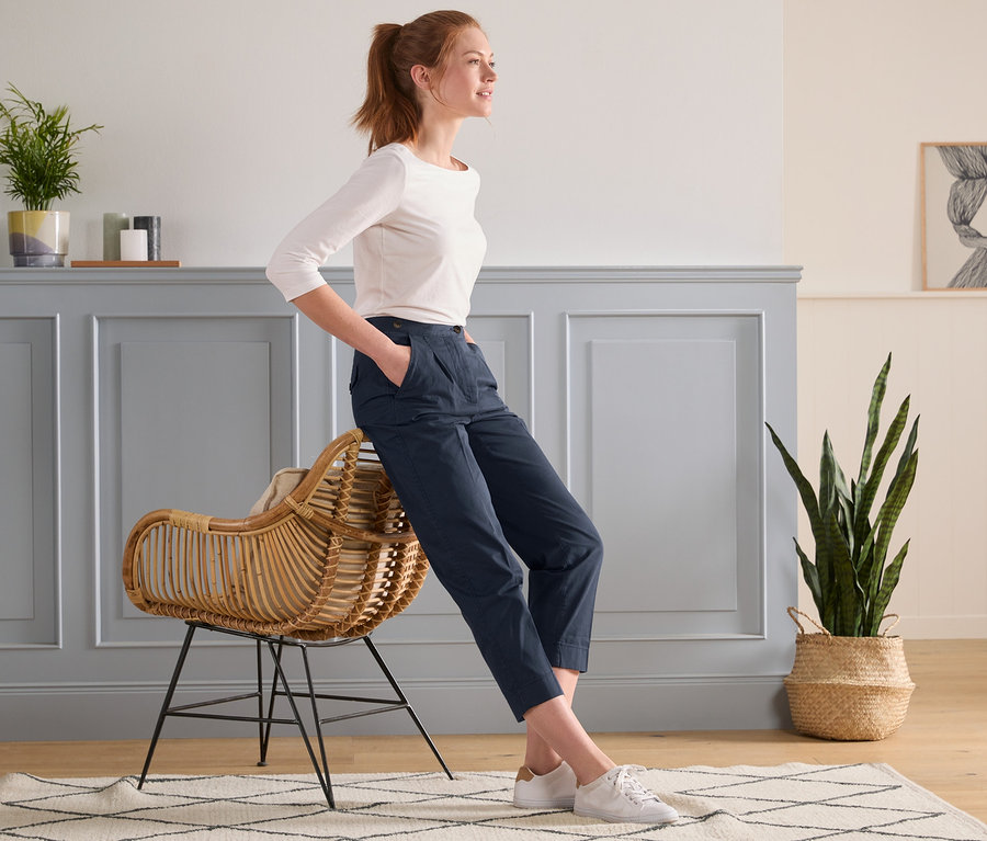 Eine Frau lehnt an einem Rattansessel und trägt eine moderne dunkelblaue Chino.