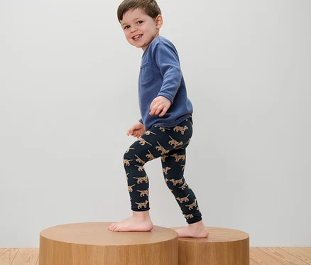 Junge mit blauem Sweatshirt und 2 Kinder-Strickleggings mit Stickerei steht auf einem Holzpodest.