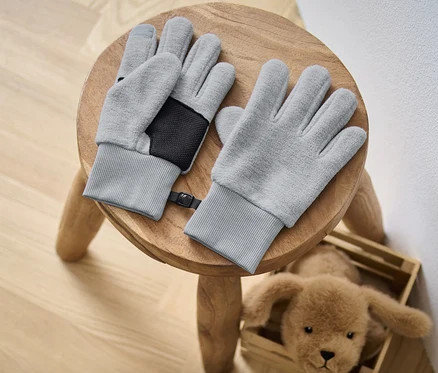 Zwei Kinder-Fleecehandschuhe liegen auf einem Holzhocker neben einem Plüschhund.