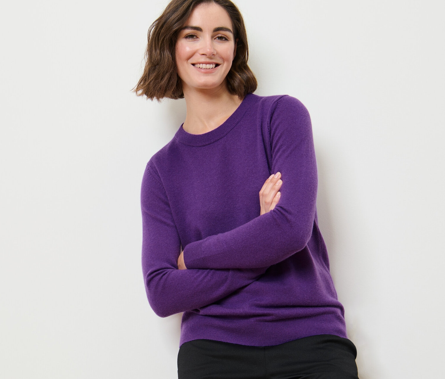 Frau posiert mit verschränkten Armen in einem lila Feinstrickpullover mit Cashmere und einer schwarzen Hose.