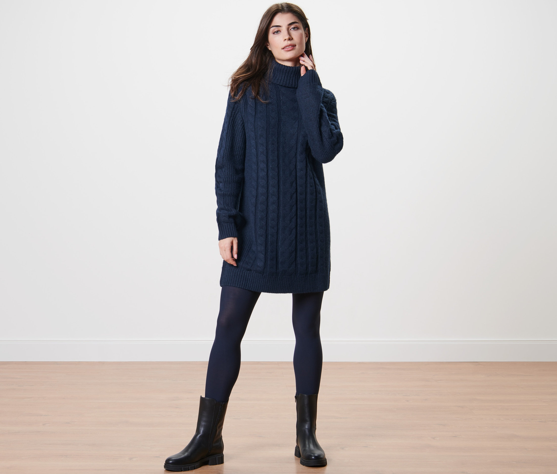 Frau posiert in langem Strickpullover mit Zopfmuster, dunkler Strumpfhose und schwarzen Stiefeln.