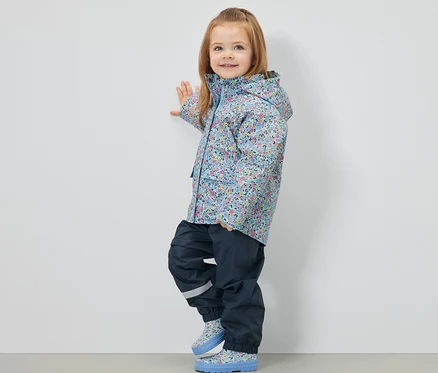 Mädchen steht seitlich und trägt eine geblümte Kinder-Regenjacke, dunkelblaue Hosen und Stiefel.
