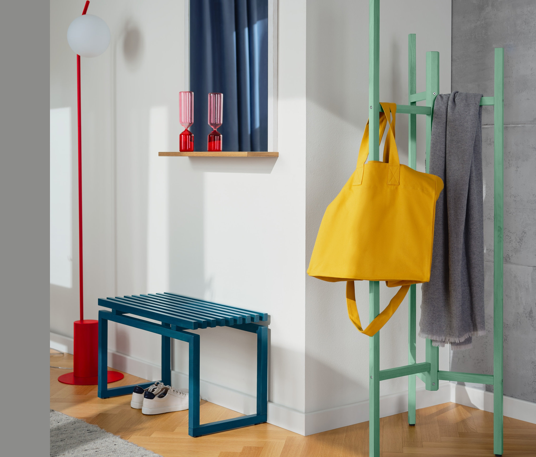 Eine blaue Bank steht an einer Wand. An der Wand hängt ein Wandspiegel mit Ablage. Neben dem Spiegel steht ein Garderobenständer, an dem eine gelbe Tasche und ein grauer Schal hängen.