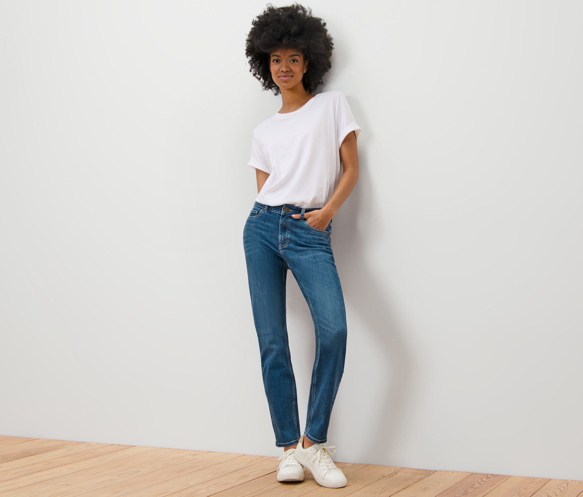 Eine Frau posiert an einer Wand, sie trägt eine Slim Jeans – Fit »Emma«, mid blue, ein weißes T-Shirt und Turnschuhe.