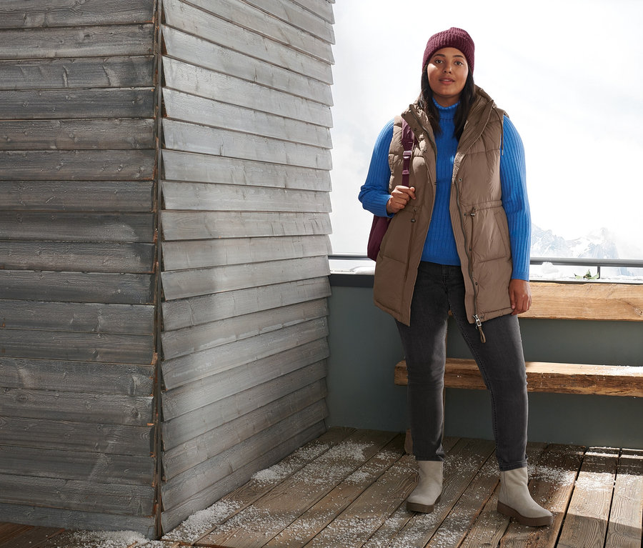 Eine Frau steht draußen und trägt eine Steppweste, einen gerippten Rollkragenpullover, Slimfit-Jeans, Veloursleder-Boots und eine bordeauxrote Strickmütze. Sie trägt einen gesteppten Outdoor-Rucksack.