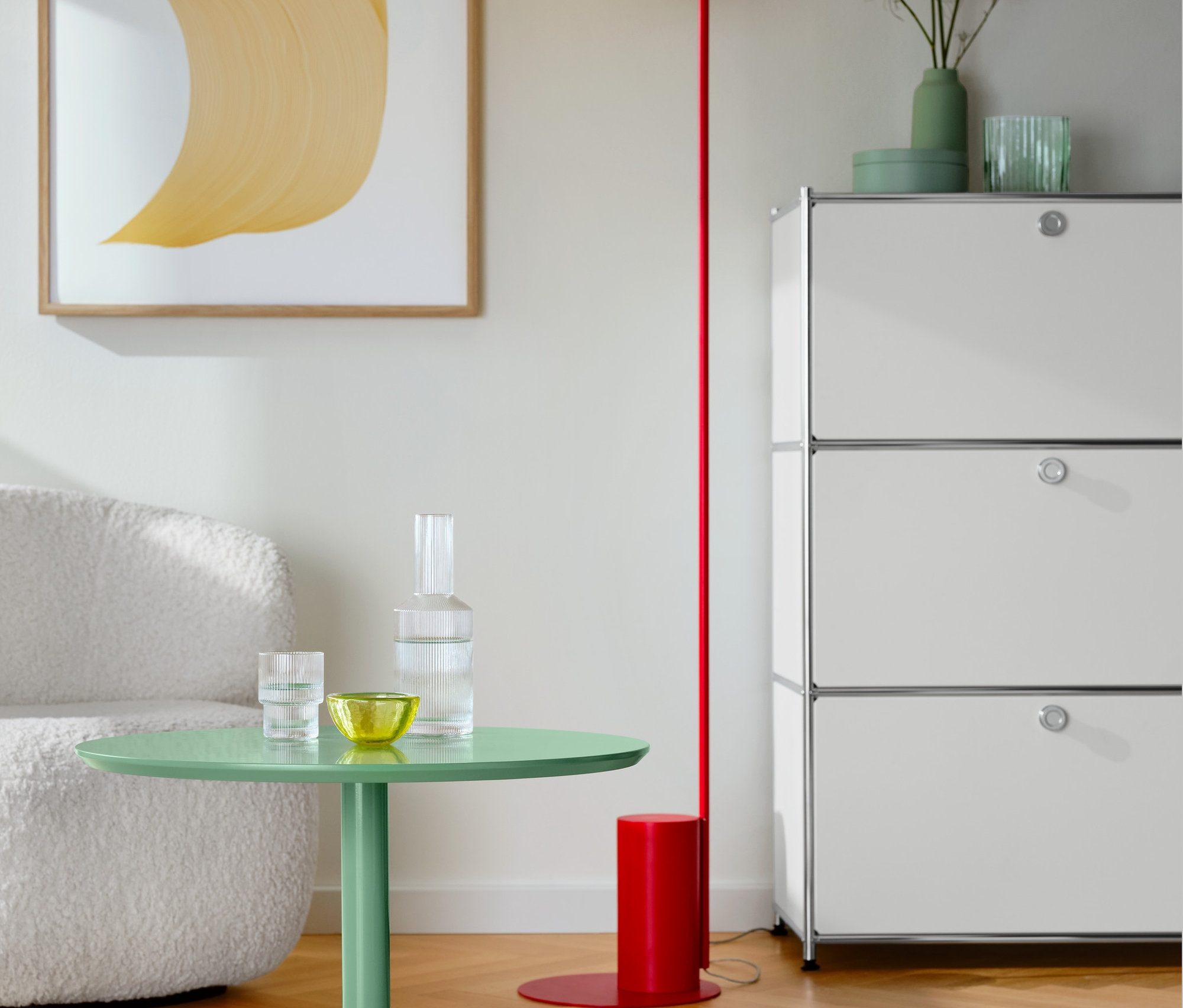 Im Wohnzimmer stehen ein runder Couchtisch in Salbei, ein Regal Metall »CN3« mit 3 Klappenfächern in Weiß, ein Sessel mit Drehfunktion und eine rote Stehleuchte mit Glaskugel.
