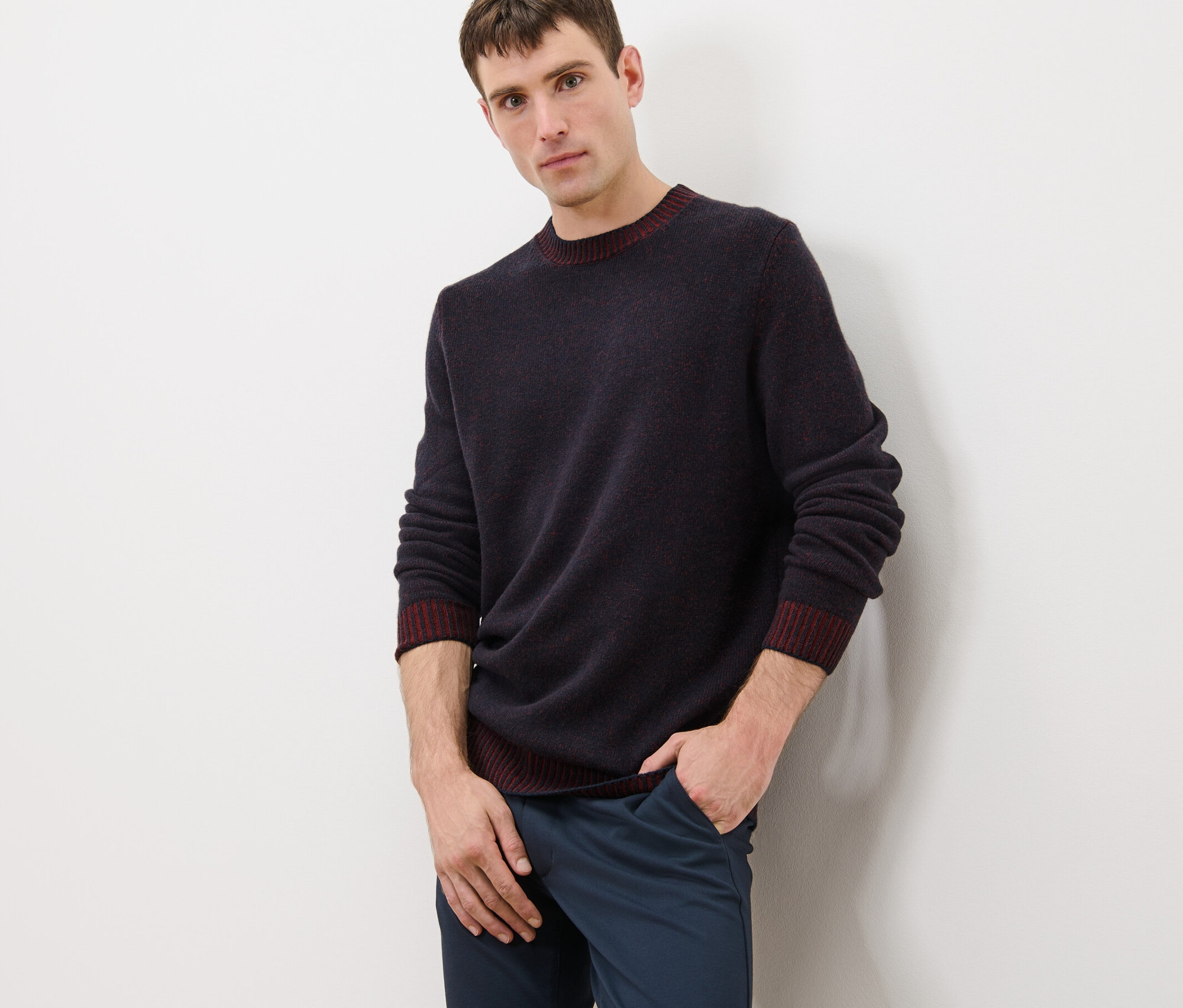 Mann posiert in dunkelblauem Pullover mit Wolle und weinrotem Saum und blauen Hosen.