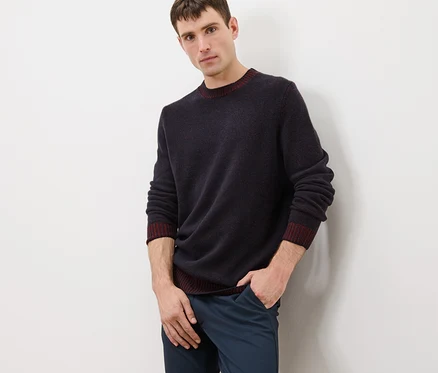 Mann posiert in dunkelblauem Pullover mit Wolle und weinrotem Saum und blauen Hosen.