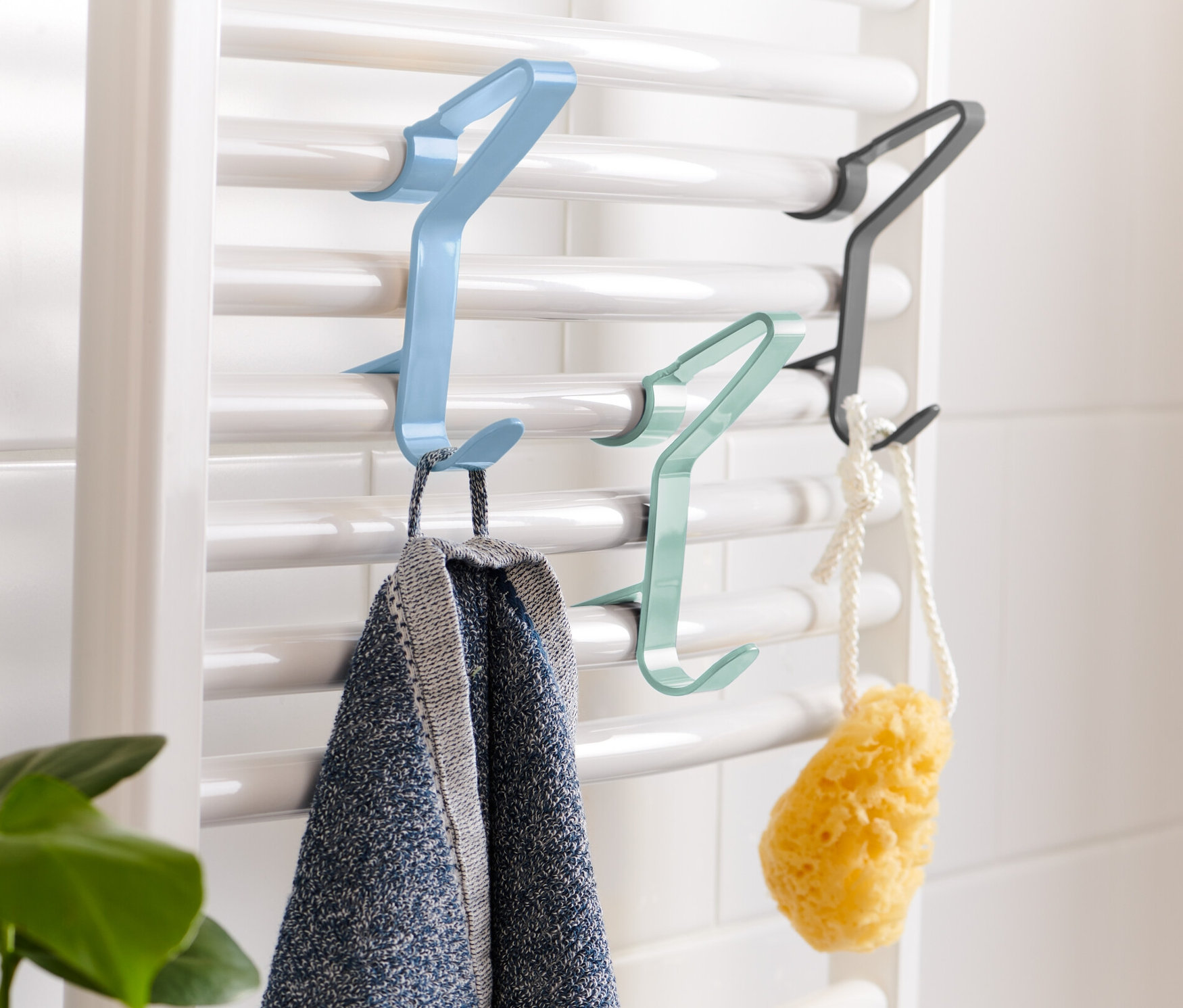 An einer weißen Heizung hängen blaue, grüne und schwarze 3 Heizkörper-Haken. An dem blauen Haken hängt ein Handtuch.