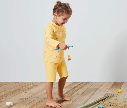 Mädchen spielt auf einer Decke mit Sand und gelbem Kinder-Kombi mit UV-Schutzfaktor 80.