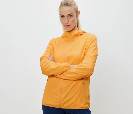 Frau posiert mit verschränkten Armen in orangefarbener, packbarer Funktionsjacke.