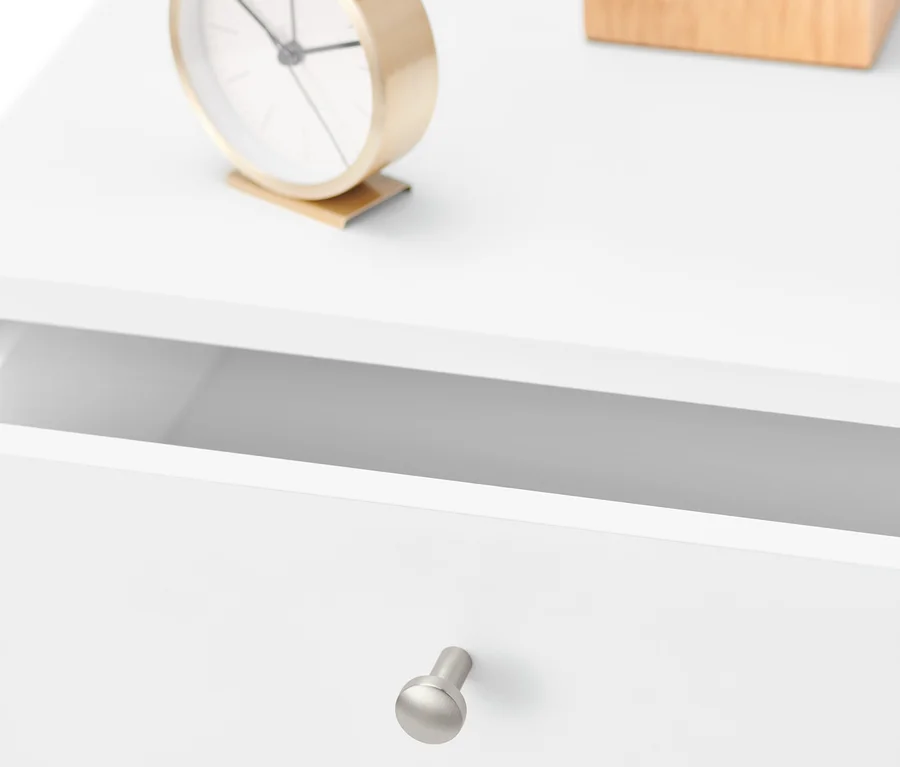 Detailansicht eines weißen Nachttisches mit offener Schublade, goldener Uhr und einer Holzkiste.