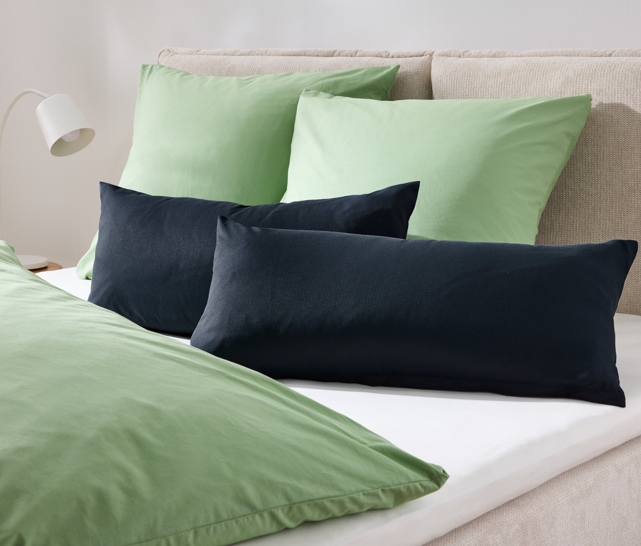 Auf einem Bett liegen zwei grüne Kissen und zwei dunkelblaue Kissen, bedeckt mit grüner Bettwäsche.