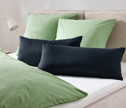 Auf einem Bett liegen zwei grüne Kissen und zwei dunkelblaue Kissen, bedeckt mit grüner Bettwäsche.