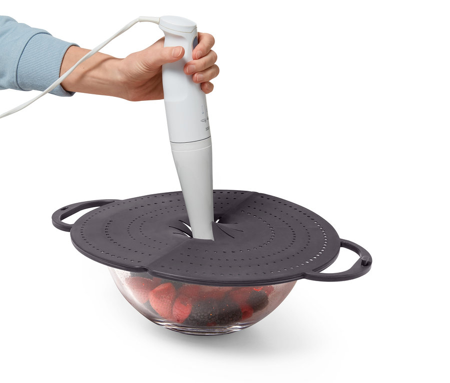 Eine Hand hält einen weißen Stabmixer über eine mit Obst gefüllte Schüssel, die mit einem grauen Deckel mit Löchern abgedeckt ist.