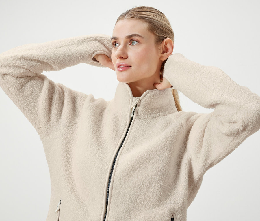 Frau mit blonden Haaren, die zu einem Pferdeschwanz zusammengebunden sind, posiert in einer cremefarbenen Fleecejacke mit Reißverschluss.