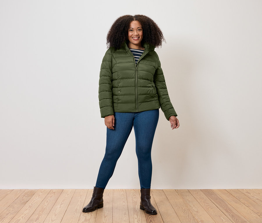 Eine Frau steht und trägt eine olivgrüne Steppjacke, Jeans und braune Stiefel.