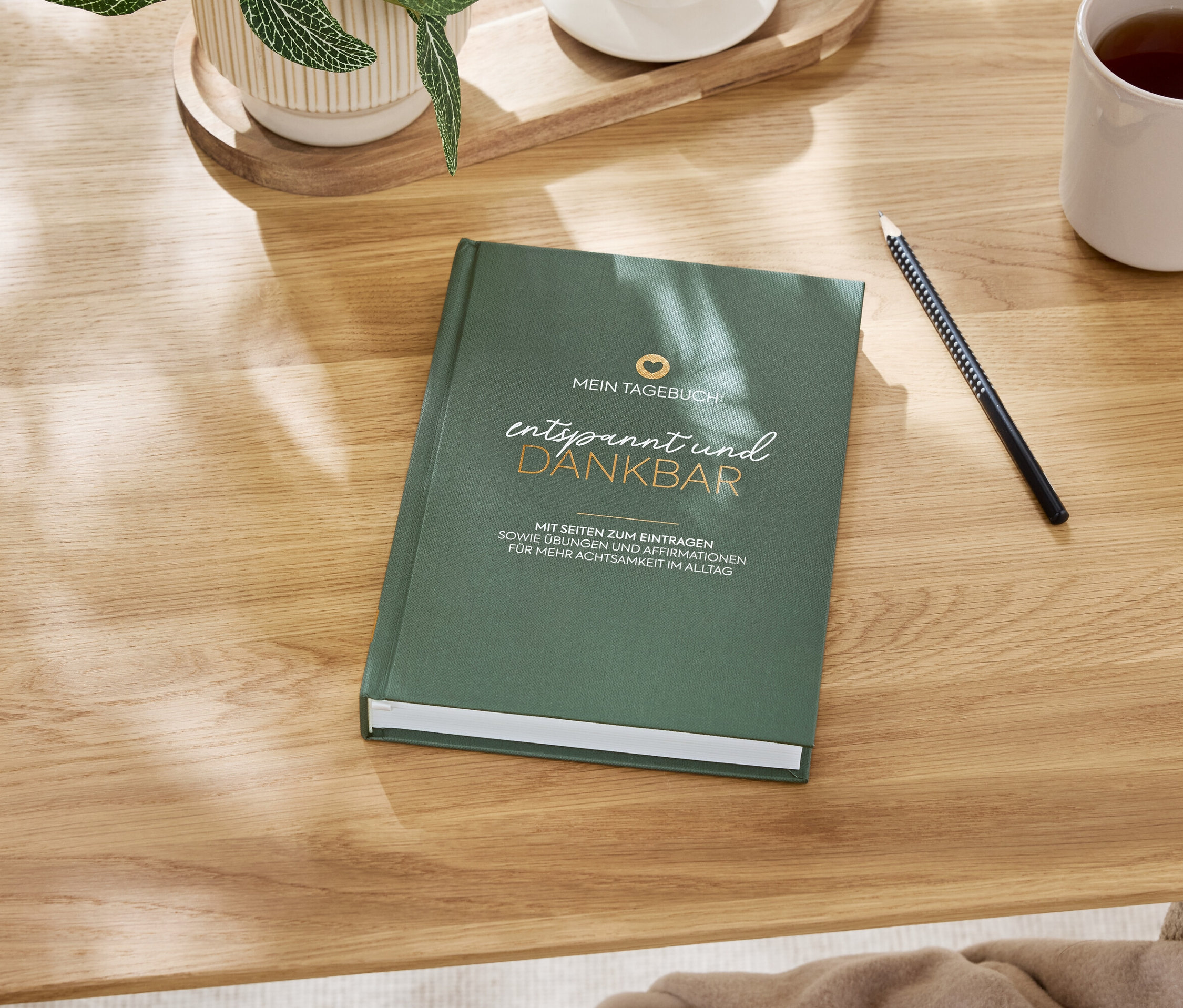 Ein grünes Notizbuch 'Entspannt und dankbar' liegt auf einem Holztisch neben einem Stift und einer Tasse mit Getränk.