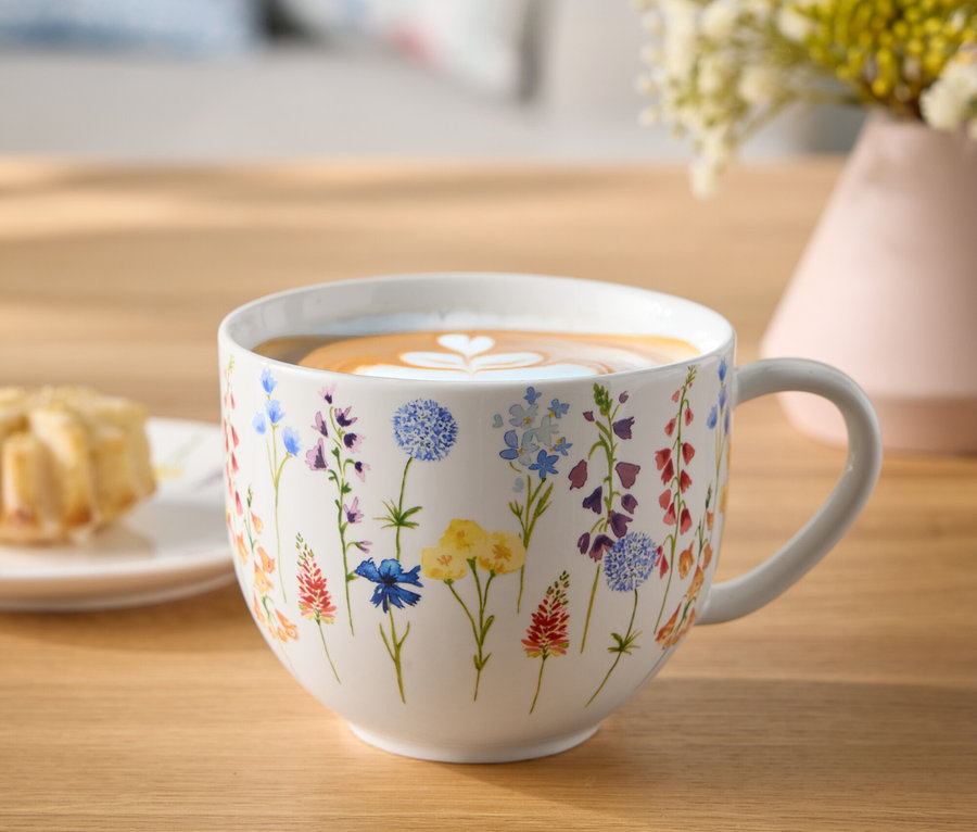 Blumenbecher mit Kaffee und Latte-Art-Herz auf einem Holztisch neben einem Teller mit Gebäck und einer Blumenvase.