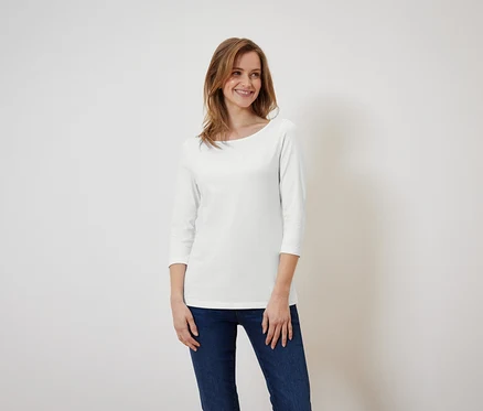 Eine lächelnde Frau posiert vor einem weißen Hintergrund in einem weißen Dreiviertelarm-Shirt und blauen Jeans.