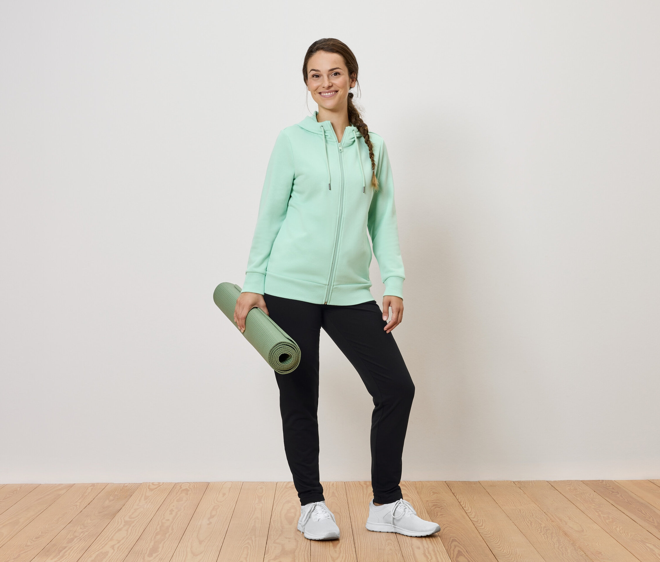 Frau hält eine zusammengerollte grüne Yogamatte, trägt einen mintfarbenen Kapuzenpullover und eine schwarze Trainingshose.