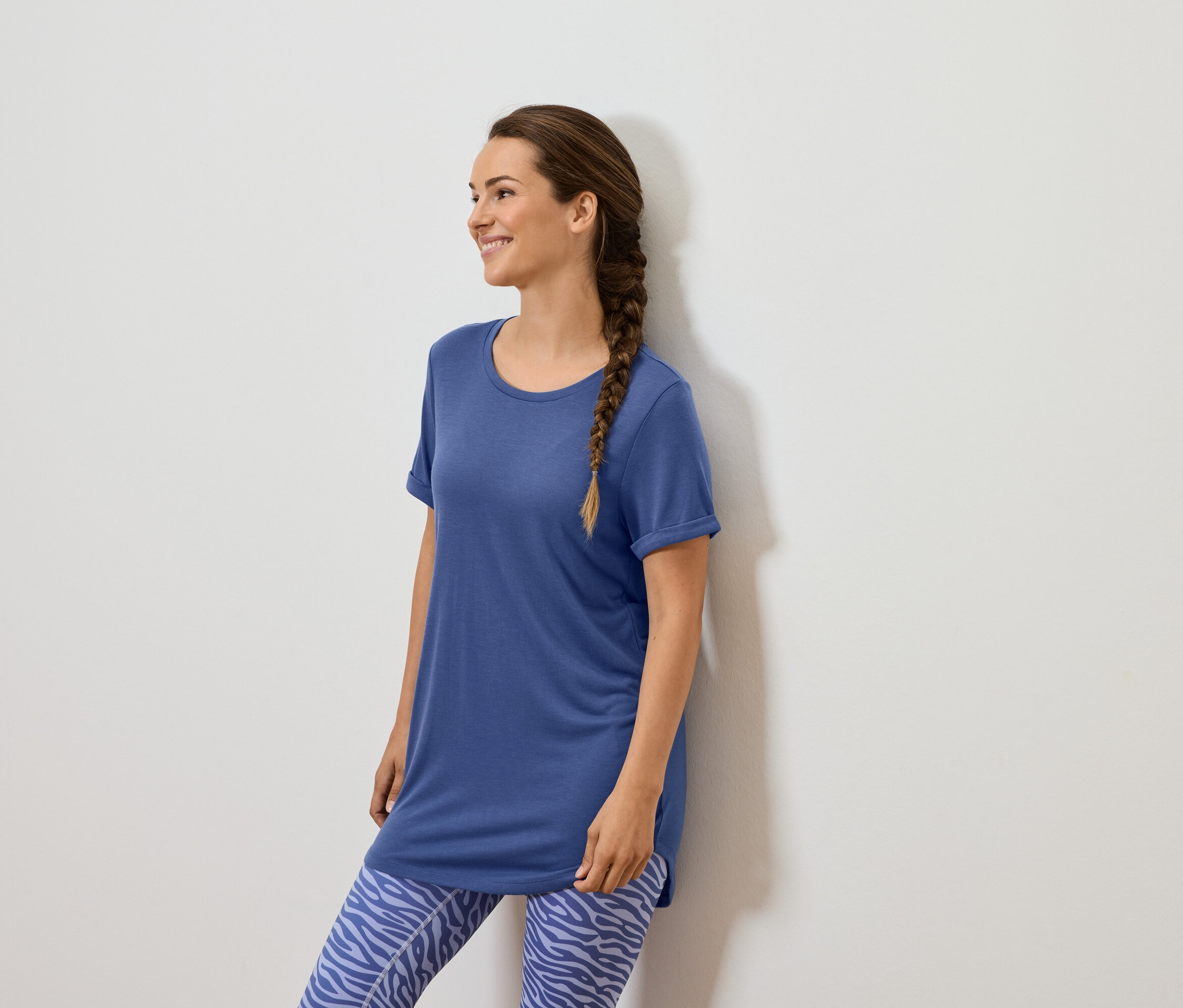 Eine Frau mit braunem Zopf steht an einer weißen Wand und trägt ein blaues T-Shirt und blaue Leggings mit Tiermuster.