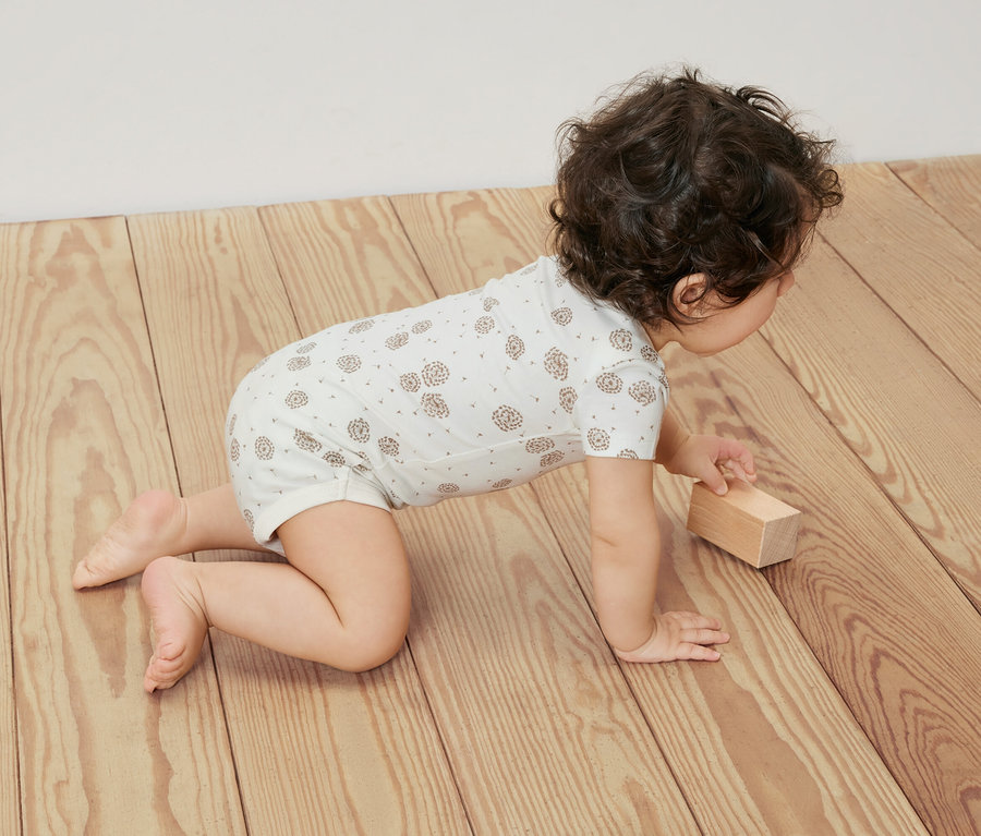 Baby krabbelt auf einem Holzboden und trägt einen weißen 5er-Pack Baby-Bodys mit Mitwachsfunktion.