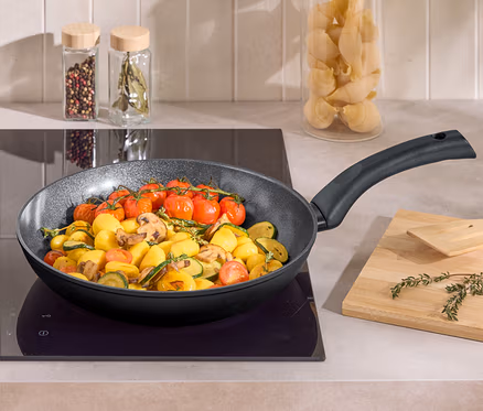 In einer Bratpfanne auf einem Kochfeld werden Kartoffeln, Zucchini, Champignons und Cherrytomaten gebraten. Daneben liegen ein Holzbrett und ein Pfannenwender.