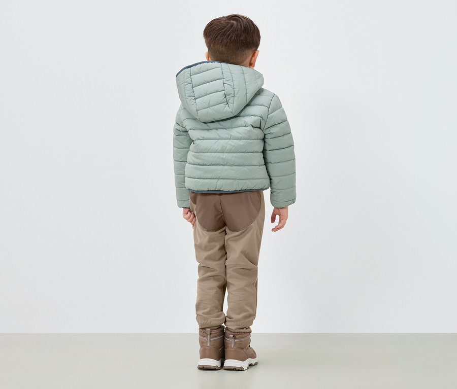 Ein Junge steht mit dem Rücken zum Betrachter und trägt eine hellgrüne Daunenjacke mit Kapuze, braune Hosen und braune Stiefel.