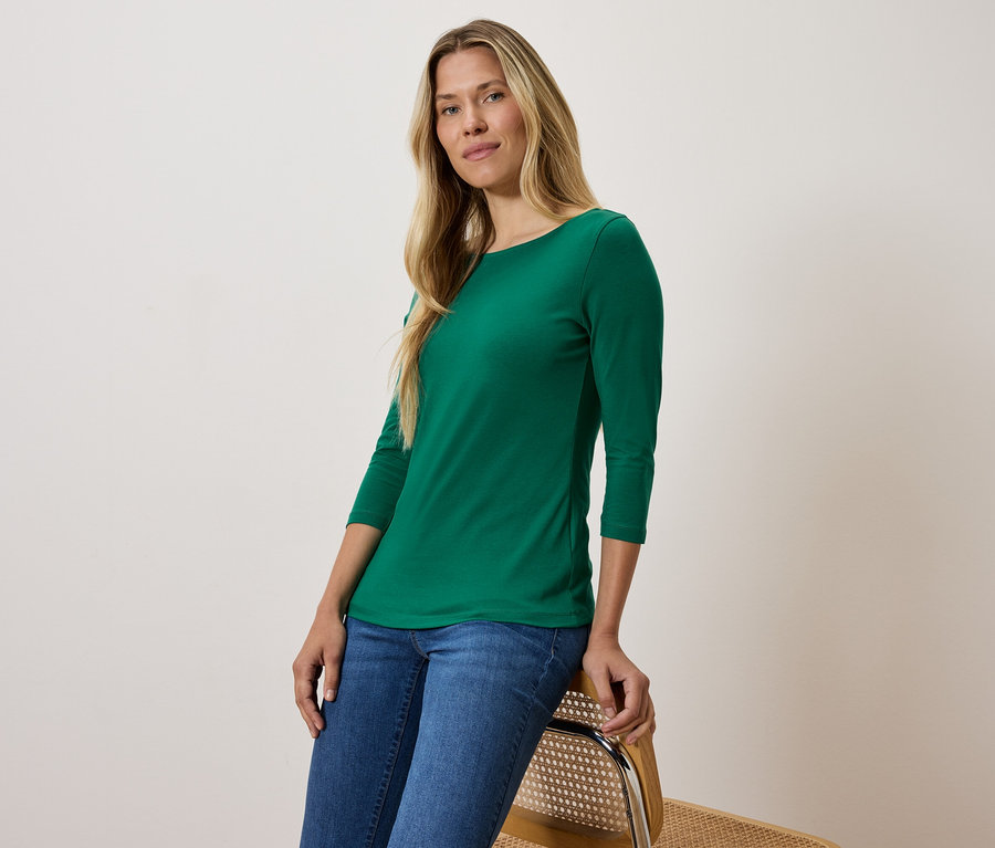 Blonde Frau in grünem Dreiviertelarm-Shirt und blauen Jeans sitzt auf einem Stuhl.