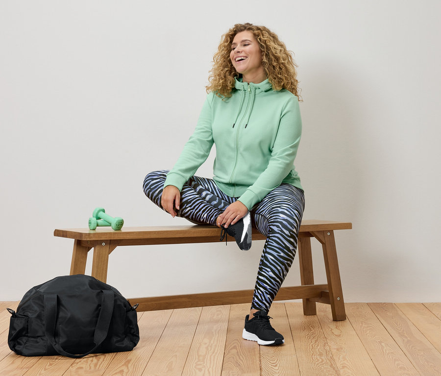 Frau mit lockigen Haaren sitzt auf einer Holzbank, trägt einen mintgrünen Hoodie und Leggings mit Zebramuster. Eine schwarze Sporttasche und grüne Hanteln sind ebenfalls sichtbar.