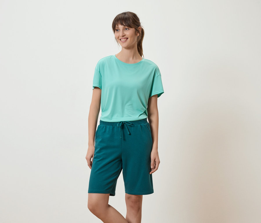 Frau mit hellgrünem T-Shirt und grünen Shorts steht vor einem weißen Hintergrund.