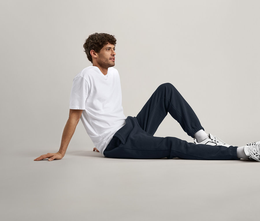 Mann mit lockigem Haar sitzt auf dem Boden, trägt ein weißes T-Shirt, marineblaue Jogginghose und Turnschuhe.