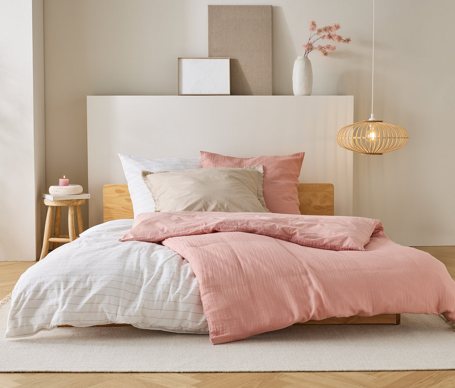Ein Bett mit rosa Bettdecke, weißen Laken und beigen Kissen steht auf einem Teppich in einem Schlafzimmer.