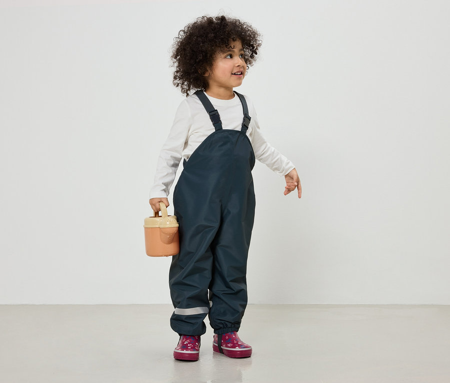 Ein Kind mit lockigem Haar trägt eine dunkelblaue Latzhose und rosa Stiefel und hält eine kleine Gießkanne.