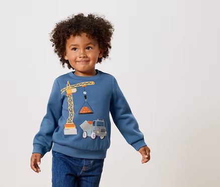 Ein lockiger Junge trägt ein blaues Sweatshirt mit einer gelben Kran- und Lastwagen-Applikation.
