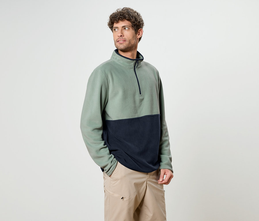 Ein Mann mit lockigem Haar trägt ein zweifarbiges Fleece-Oberteil in Grün und Marineblau mit beigen Shorts.