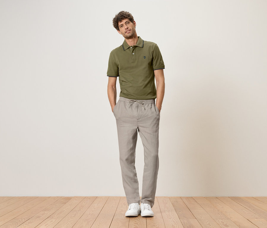 Mann steht mit Händen in den Hosentaschen, trägt ein grünes Poloshirt, graue Leinenhose und weiße Turnschuhe.