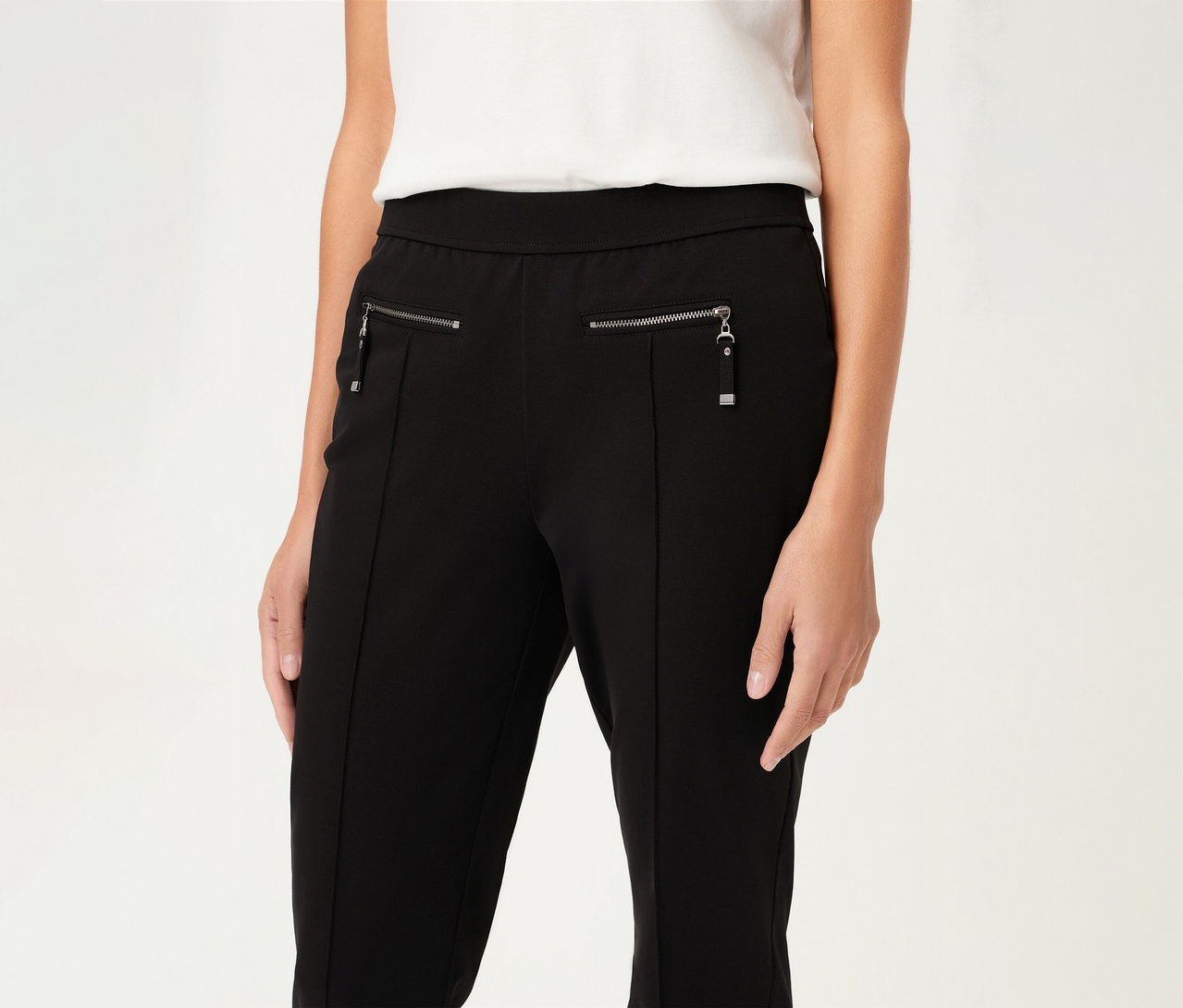 Frau trägt eine Olsen Hose, slim, in Schwarz mit Reißverschlüssen. Sie trägt ein weißes Oberteil.