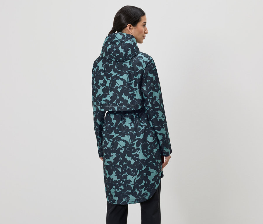 Frau von hinten, trägt einen blaugrünen, langen Regenmantel mit Kapuze und schwarzem Blumenmuster, dazu schwarze Hose.